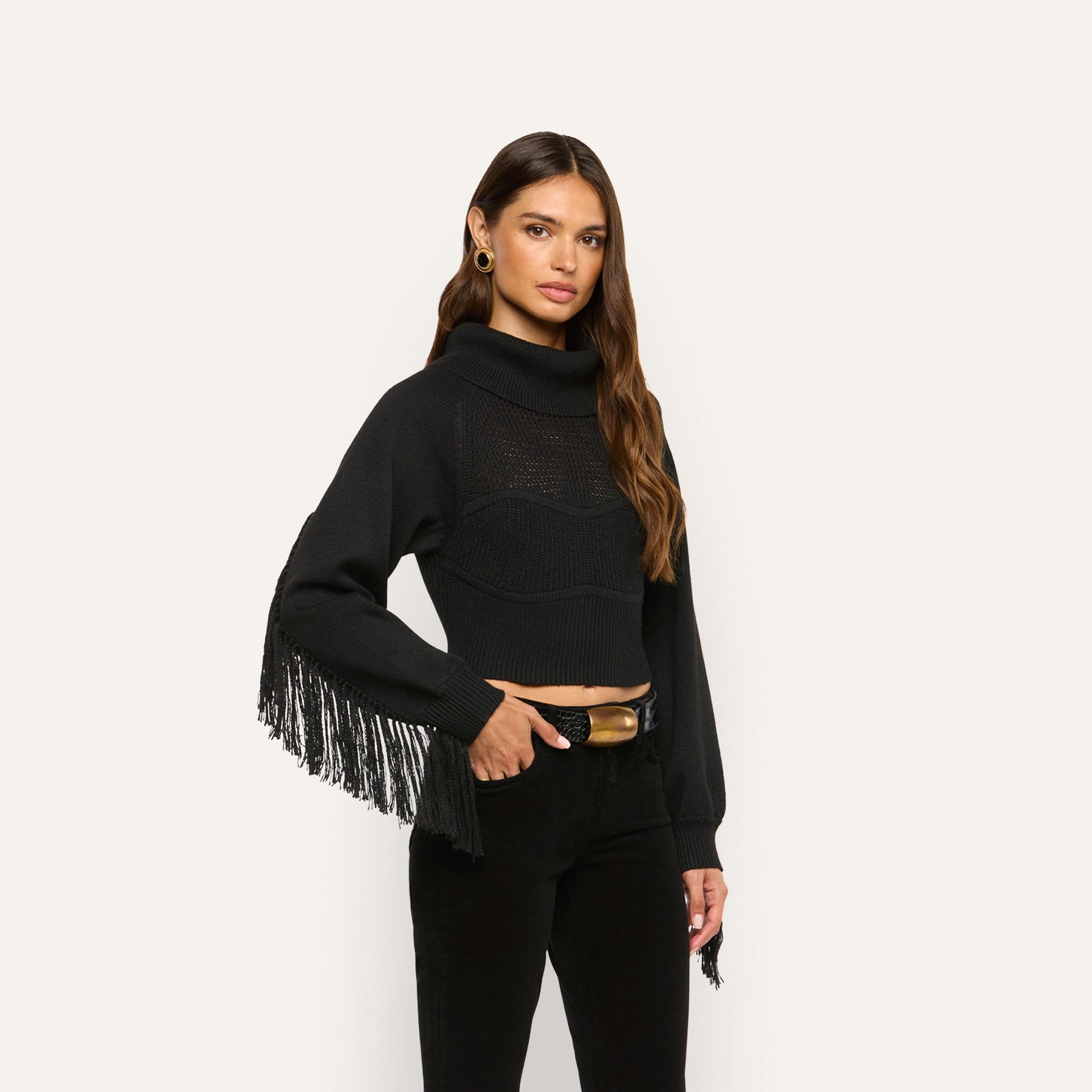 A woman wears a marled black Maddison Turtleneck featuring a decorative knit pattern across the chest and long fringe along the sleeves, paired with black jeans and a gold-buckled belt.