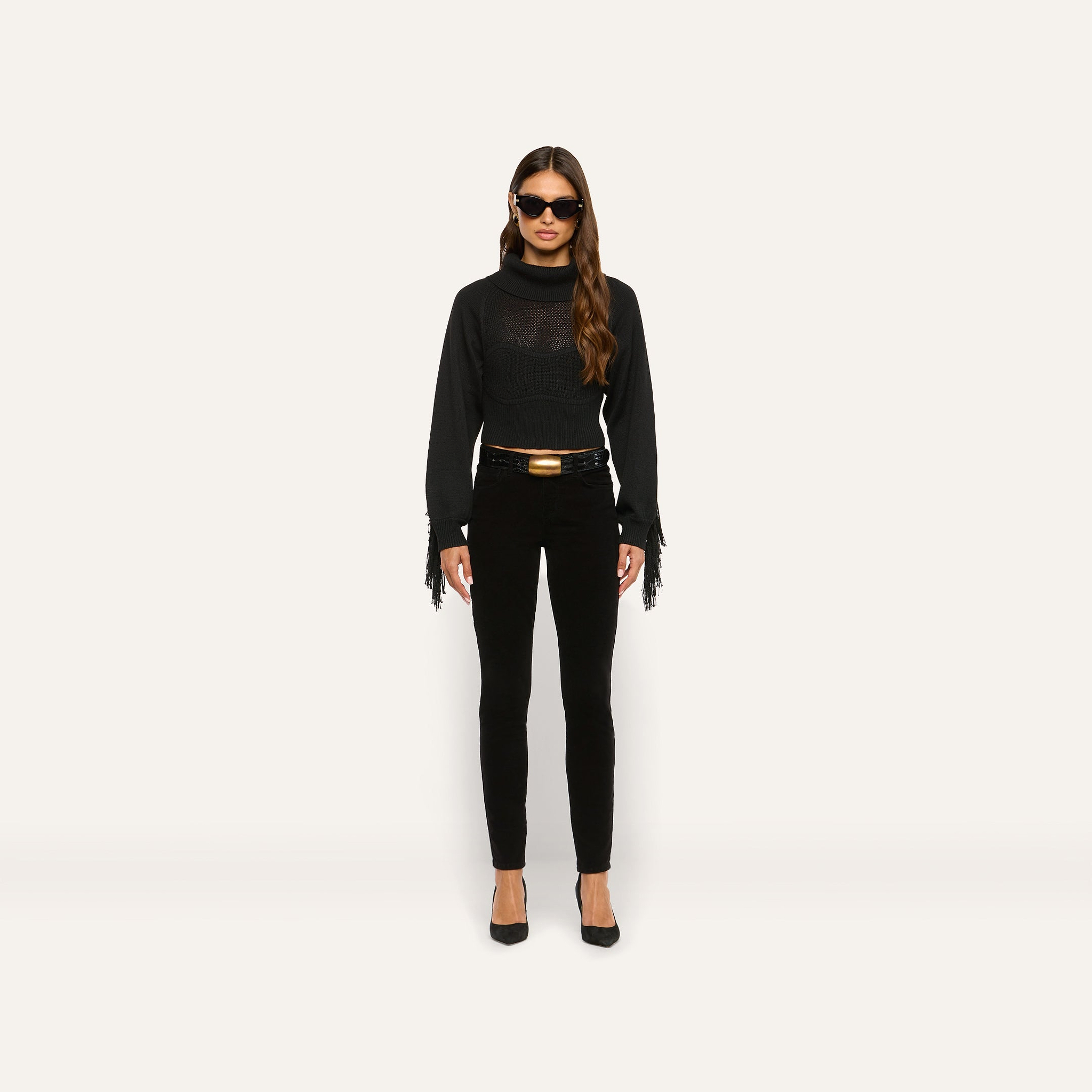 A person wears a marled black Maddison Turtleneck with a cropped turtleneck, fringe sleeves, and a corset-style bodice, paired with slim-fit pants, a belt with a gold buckle, and black heels.