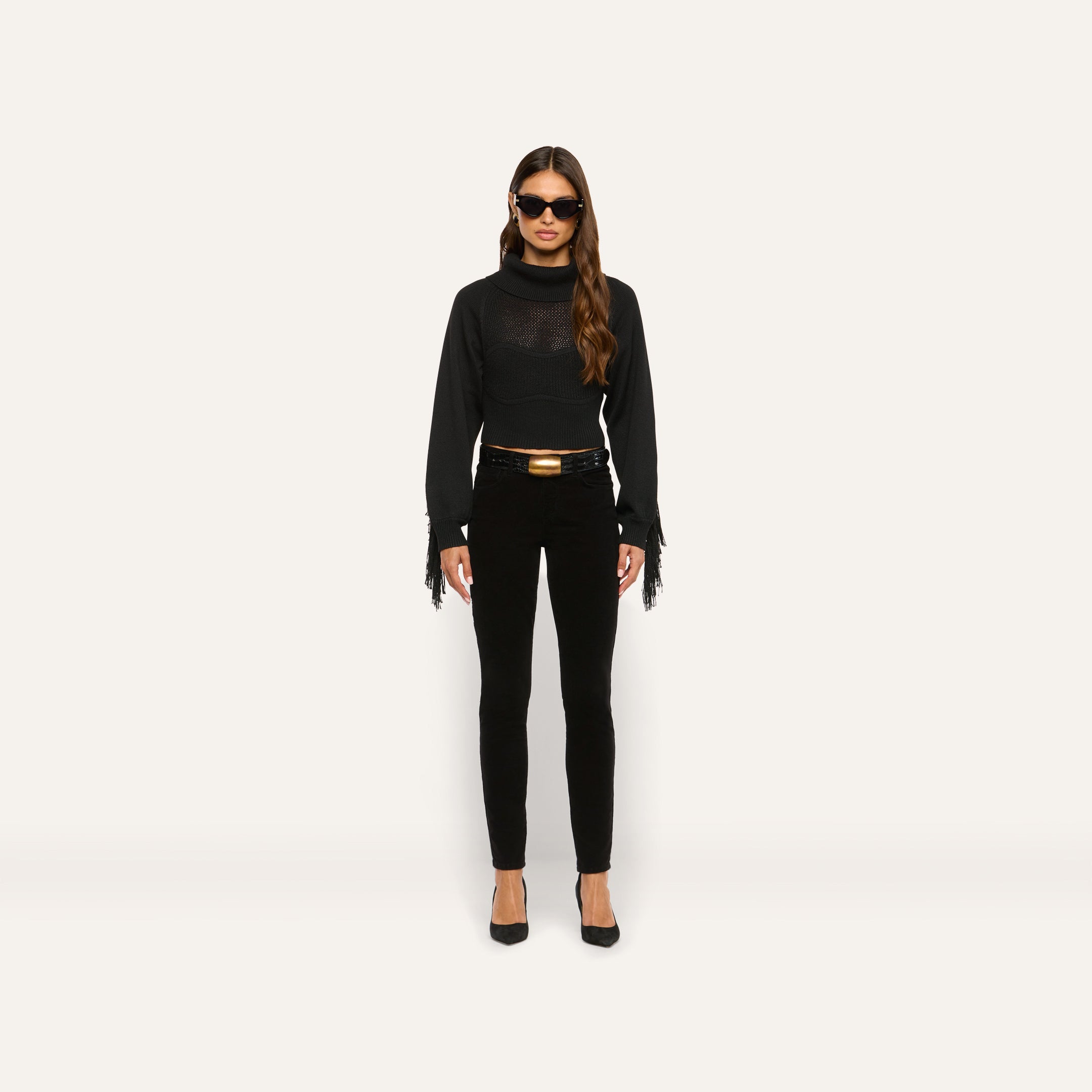 A person wears a marled black Maddison Turtleneck with a cropped turtleneck, fringe sleeves, and a corset-style bodice, paired with slim-fit pants, a belt with a gold buckle, and black heels.