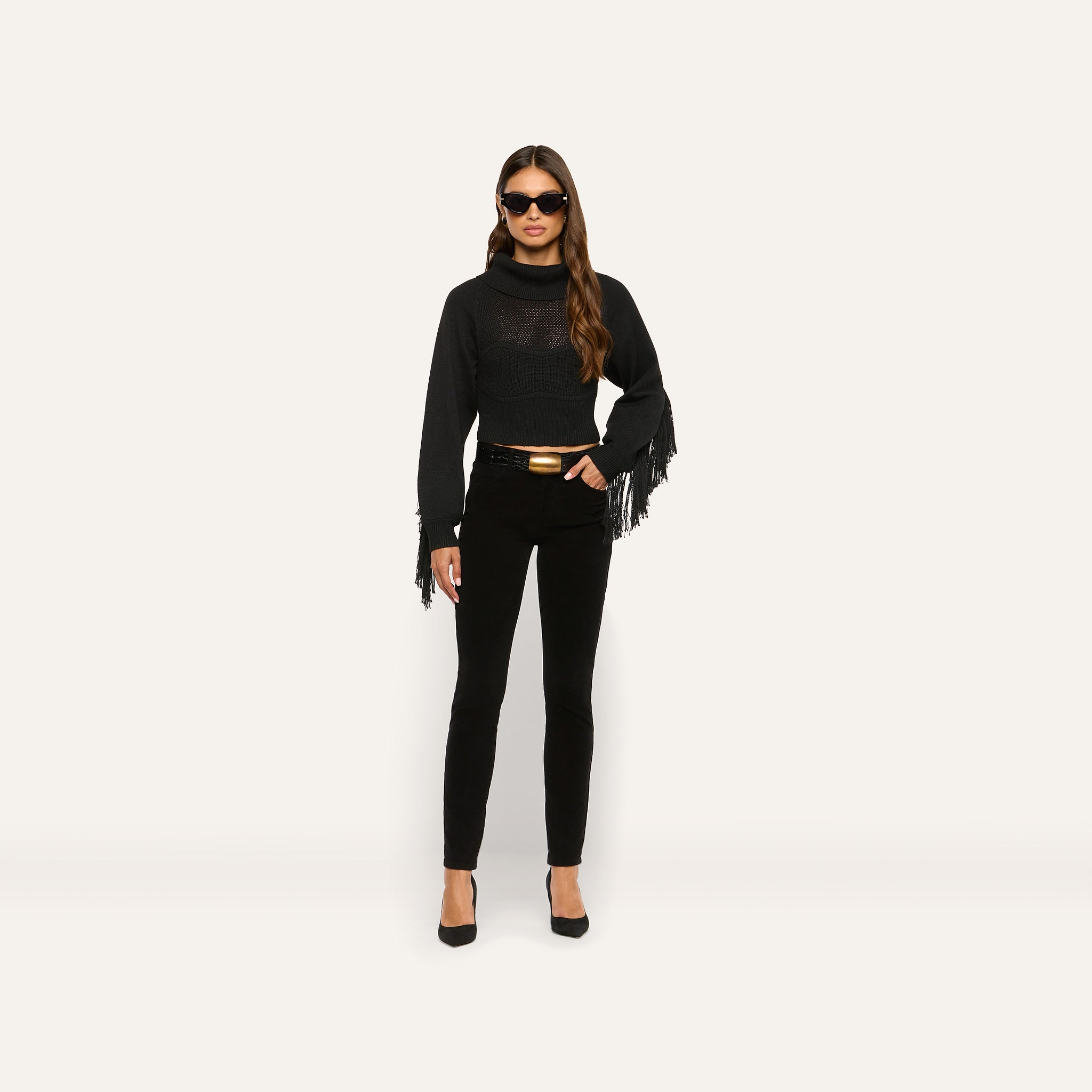 A person wearing a black Maddison Turtleneck with fringe on the sleeves, black pants with a gold-buckle belt, and black heels.