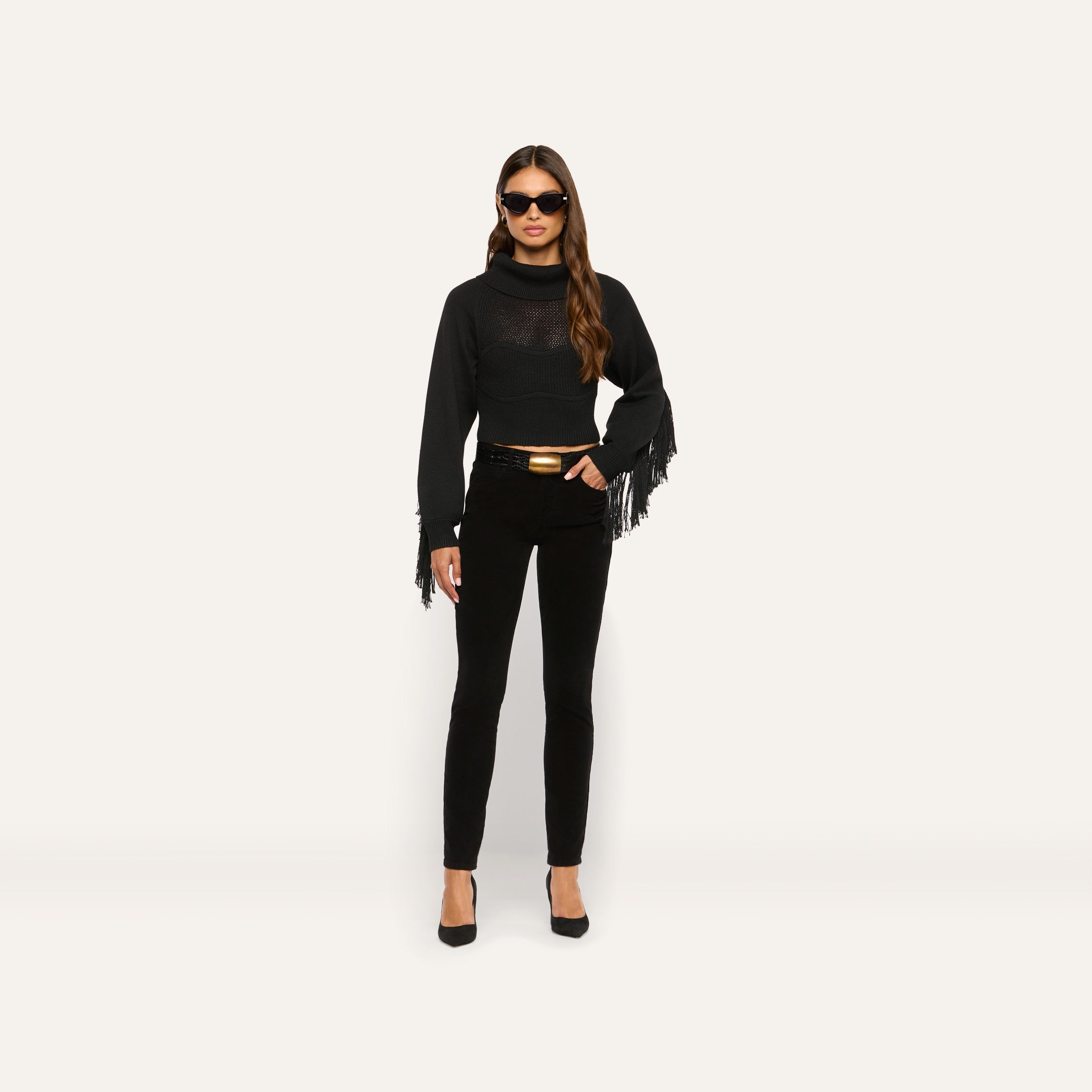 A person wearing a black Maddison Turtleneck with fringe on the sleeves, black pants with a gold-buckle belt, and black heels.