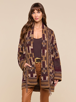 A woman with long brown hair is wearing the Jair Cardigan, featuring a patterned brown and tan design in a boho-chic style. She has paired it with a brown top and tan pants, and stands against a beige background with a neutral expression.