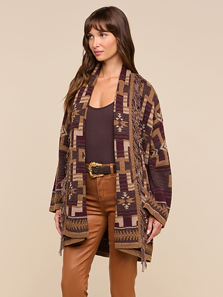 A woman stands against a beige background wearing the Jair Cardigan—a boho-chic, patterned maroon and tan shawl—layered over a brown top, paired with tan pants and a decorative belt.