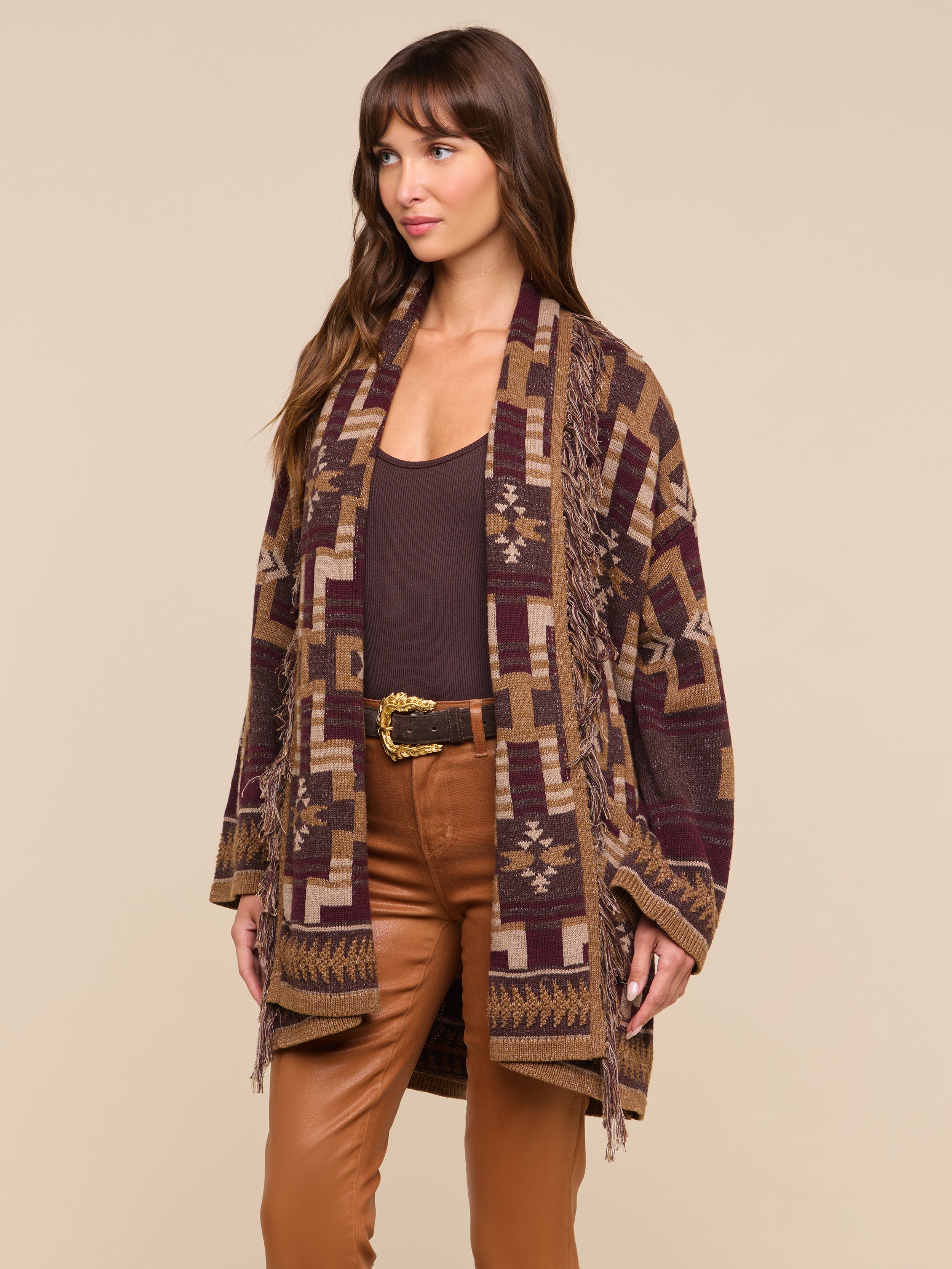 A woman stands facing slightly to the left against a plain light background, wearing the Jair Cardigan—a patterned brown and tan shawl cardigan—layered over a dark top and tan pants.
