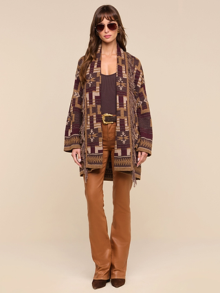 A woman wearing sunglasses, a boho-chic Jair Cardigan featuring geometric patterns, a dark top, tan flared pants, and brown boots stands facing forward against a plain beige background.