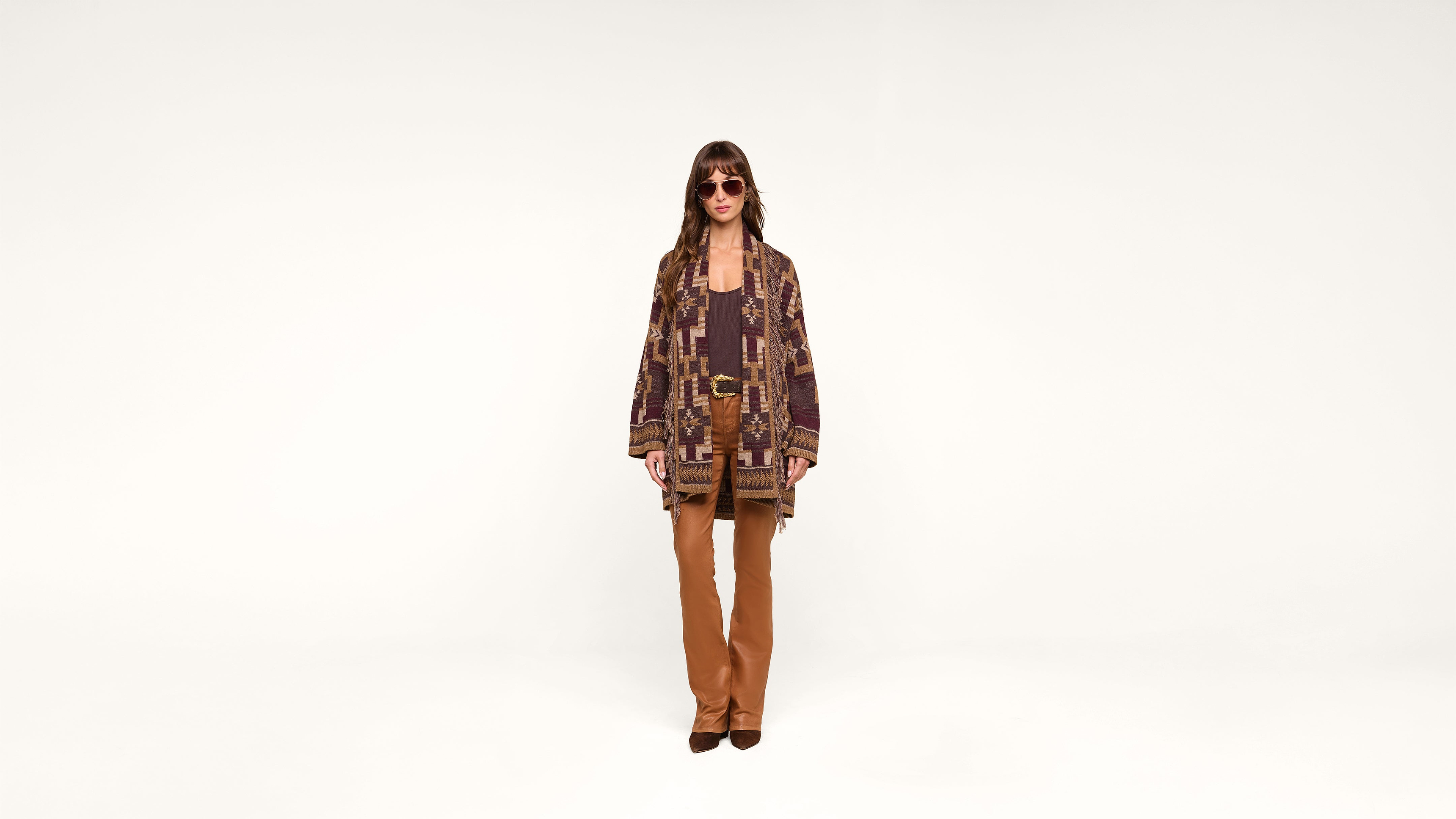 A person wearing sunglasses, a patterned brown and purple cardigan, brown top, and tan pants stands against a plain white background, facing forward.
