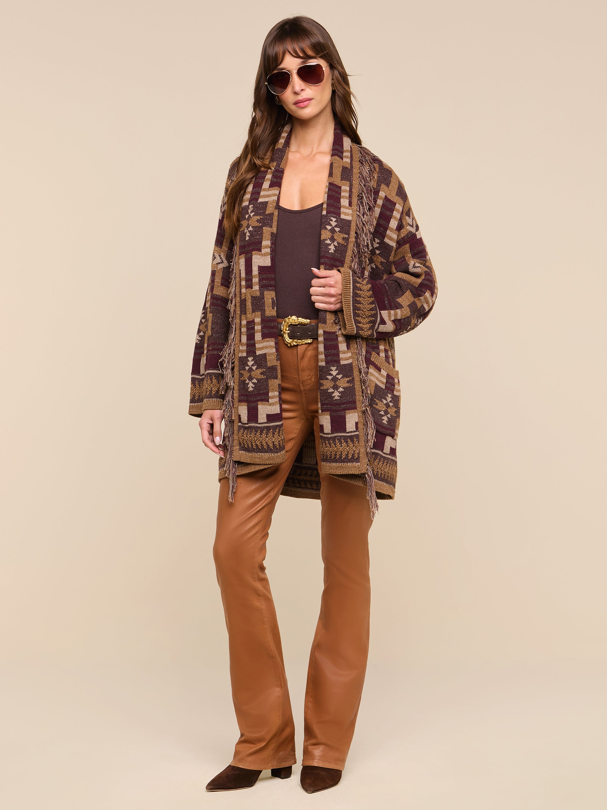 A woman wearing sunglasses, the Jair Cardigan in a boho-chic style, a brown top, brown flared pants, and brown shoes stands confidently against a plain white background.