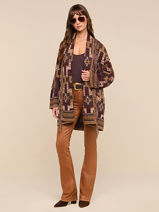 A woman wearing sunglasses, a boho-chic Jair Cardigan in a patterned brown design, a dark brown top, tan leather flared pants, a belt with a gold buckle, and brown heeled boots stands against a beige background.