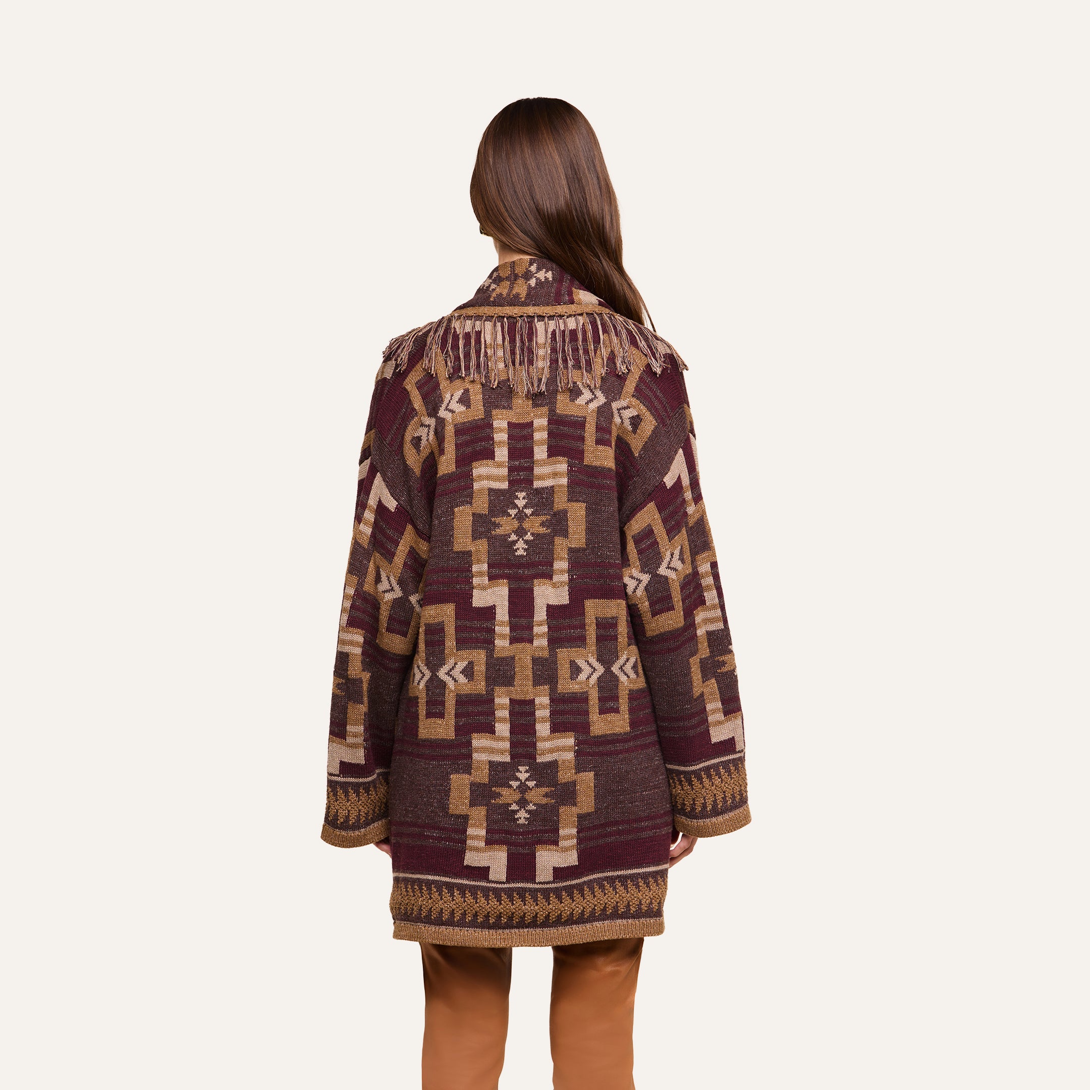 A person with long brown hair stands with their back to the camera against a plain background, wearing the Jair Cardigan in maroon and tan with a patterned design and fringe detailing across the upper back.