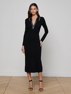 A woman stands indoors on a wooden floor wearing the L'AGENCE Sale Yaretzi Knit Midi Dress, a fitted black midi with long sleeves and a zippered collared neckline, paired with black heels against a plain light gray background.