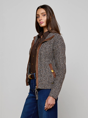 A woman with long brown hair wears the Shiloh Cable Knit Cardigan by L'AGENCE, paired with a dark brown button-up shirt, blue jeans, and a belt, standing against a plain light gray background.