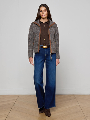 A woman with long straight hair poses indoors against a light-colored wall, wearing the L'AGENCE Shiloh Cable Knit Cardigan over a brown button-up shirt, blue wide-leg jeans, a belt, and brown boots.