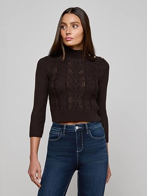 A woman with long brown hair models the L'AGENCE Kamdyn Cropped Sweater, a dark brown cable knit pullover with gold shoulder buttons, paired with high-waisted blue jeans. She stands before a plain light gray backdrop, gazing slightly left.