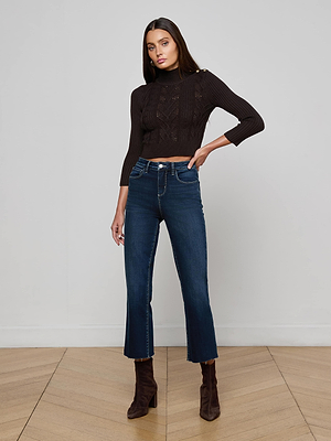 A woman with long dark hair poses confidently indoors wearing the L'AGENCE Kamdyn Cropped Sweater—a black cable knit pullover with gold shoulder buttons—paired with high-waisted flared blue jeans and brown ankle boots.