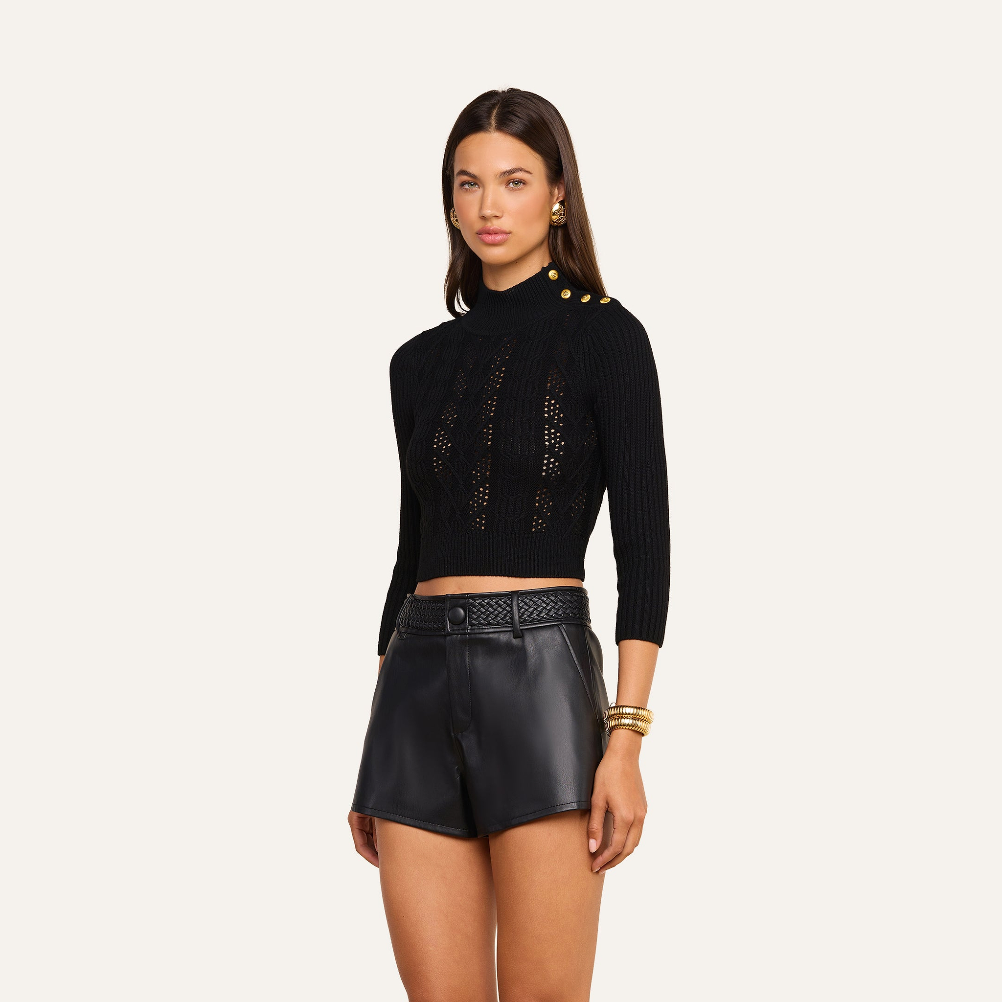 A woman wearing the Kamdyn Cropped Sweater—a black cable knit, long-sleeve crop top with gold buttons on the shoulder—paired with high-waisted black leather shorts, poses against a plain, light background.