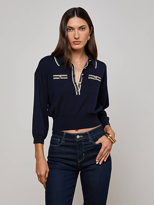 A woman with straight brown hair poses against a light gray background wearing the L'AGENCE Malaki Cropped Sweater in navy with gold-tone buttons, paired with dark blue jeans and gold jewelry. One hand rests on her hip.