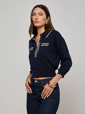A woman with long brown hair wears the L'AGENCE Malaki Cropped Sweater in navy with striped details and dark jeans. She stands against a light gray background, confidently looking at the camera with one hand on her hip.
