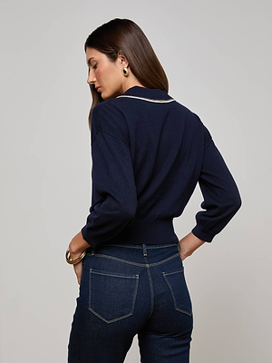 A woman with long brown hair wears the L’AGENCE Malaki Cropped Sweater, featuring light trim and gold-tone buttons, paired with dark blue jeans as she stands facing away from the camera against a plain background.
