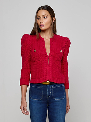 A woman with straight brown hair wears the Delli Cropped Cardigan—a bright red textured knit with gold buttons and front pockets—paired with high-waisted dark blue jeans and a gold belt, against a plain light background.