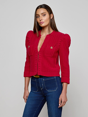 A woman with long brown hair models the Delli Cropped Cardigan, a bright red textured knit with gold buttons and puffed shoulders, paired with dark blue high-waisted jeans, standing against a plain light gray background.
