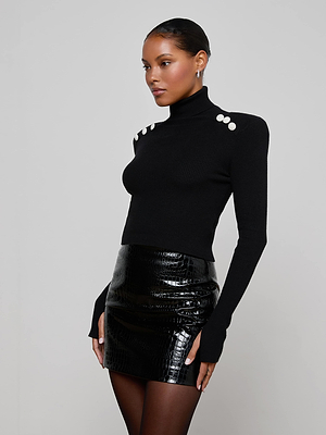A woman wears the Colette Cropped Turtleneck, a black ribbed knit sweater with statement shoulder buttons, paired with a shiny black vinyl mini skirt against a plain gray background.