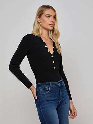 A woman with long blonde hair wears the L'AGENCE Elisabetta Knit Bodysuit, a fitted black long sleeve polo with gold buttons, paired with blue jeans. She stands against a light gray background, one hand on her hip and looking to the side.