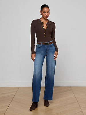 A woman stands indoors against a plain light wall, wearing the Elisabetta Knit Bodysuit in chocolate brown with gold buttons, styled with blue jeans, a black belt, and brown heeled boots. She looks at the camera with a slight smile.