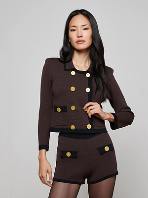 A woman with long dark hair models the L'AGENCE Janu Cropped Knit Jacket, featuring a stylish brown double-breasted design with black trim and large gold buttons, paired with matching shorts against a light gray background.