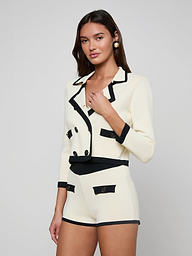 A woman wearing the Janu Cropped Knit Jacket—a cream-colored, double-breasted piece with black trim—paired with matching high-waisted shorts stands against a plain light background.