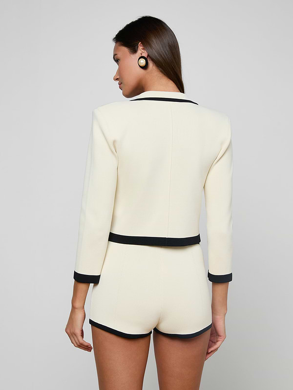 A woman stands facing away, wearing the Janu Cropped Knit Jacket in cream with black trim and matching high-waisted shorts. She has straight brown hair and wears black circular earrings against a plain light background.