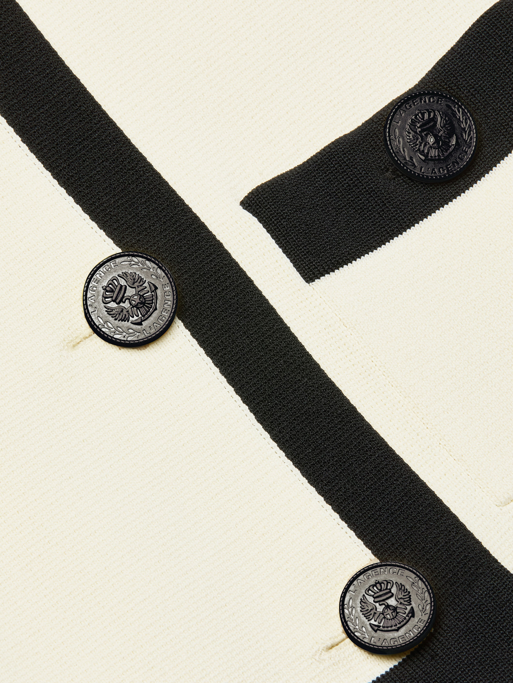 Close-up of the Janu Cropped Knit Jacket, crafted in cream-colored knit with black trim and round black embossed buttons. The double-breasted buttons showcase a crest design accented by text circling the edge. The fabric suggests this piece is a cropped cardigan-style jacket.