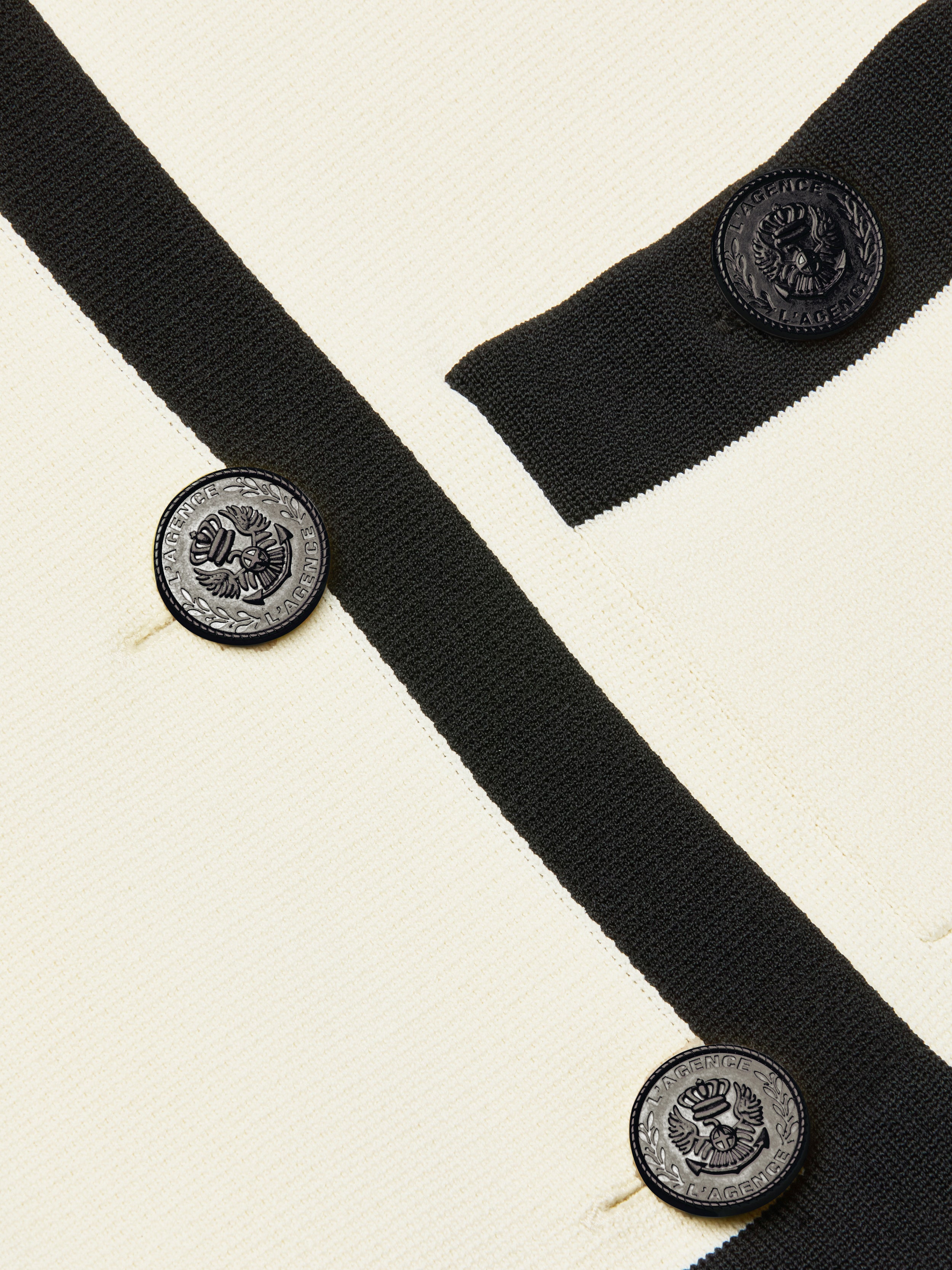 Close-up of the Janu Cropped Knit Jacket, crafted in cream-colored knit with black trim and round black embossed buttons. The double-breasted buttons showcase a crest design accented by text circling the edge. The fabric suggests this piece is a cropped cardigan-style jacket.