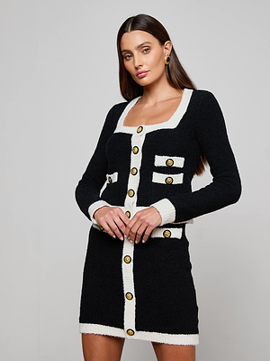 A woman with long brown hair models the Monette Cropped Cardigan by L'AGENCE, a plush black knit with a square neckline, white trim, gold buttons, and square pockets. She poses with one hand on her hip against a plain background.