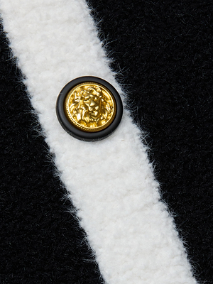 A close-up of an embossed gold button with a lion's head on plush black knit fabric, intersected by a white stripe, from the Monette Cropped Cardigan by L'AGENCE.