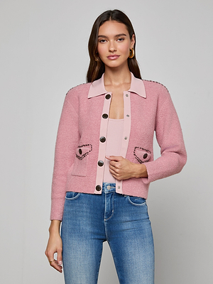 A woman with long brown hair and a neutral expression stands against a gray background, wearing the Poetry Cardigan—a light pink bouclé knit with decorative buttons and pockets—over a pink top and blue jeans.