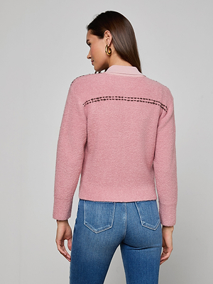 A woman with long brown hair and gold hoop earrings stands with her back to the camera, wearing the Poetry Cardigan in pink bouclé knit with a stitched horizontal pattern across the upper back, paired with blue jeans.