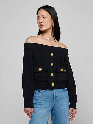 A woman wears the L'AGENCE Teri Off-The-Shoulder Top, a cropped black sweater with gold buttons and front pockets, paired with blue jeans against a plain gray background.