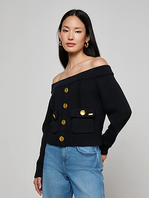 A woman with long dark hair wears the L'AGENCE Teri Off-The-Shoulder Top, a black cropped cardigan with gold buttons, paired with light blue jeans against a plain light gray background.