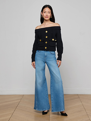 A woman poses indoors wearing the L'AGENCE Teri Off-The-Shoulder Top in black with gold buttons and front pockets, paired with wide-leg blue jeans, black heels, and gold hoop earrings on a wooden floor against a light wall.