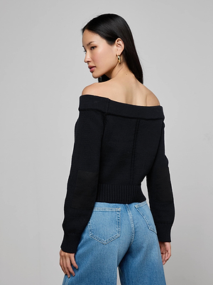 A woman with long dark hair models the L'AGENCE Teri Off-The-Shoulder Top in black, paired with blue jeans, as she looks over her shoulder against a plain light gray background.