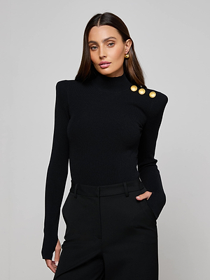 A woman with long brown hair wears the L'AGENCE Jennis Mock-Neck Bodysuit, featuring three gold shoulder buttons, paired with black pants. She stands against a plain light gray background, one hand in her pocket.