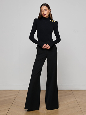 A woman with long brown hair stands indoors against a plain light wall, wearing the L'AGENCE Jennis Mock-Neck Bodysuit and wide-leg black pants, hands clasped.