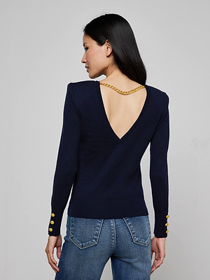 A woman with long dark hair models the Kolie Chain Top, a refined navy knit sweater with a deep V-back, high-shine chain detail, and gold button-accented sleeves. She completes the look with classic blue jeans.