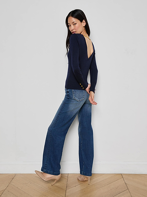 A woman stands sideways against a white wall, looking over her shoulder, wearing the Kolie Chain Top with blue jeans and nude heels.