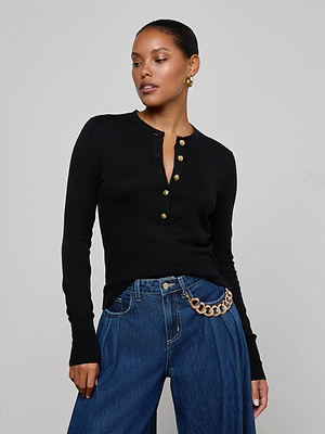 A woman models the L'AGENCE Ronda Knit Henley Top—a black lightweight knit with lion head buttons—paired with blue jeans and a gold chain belt. She wears hoop earrings, her hair pulled back, against a plain light gray background.