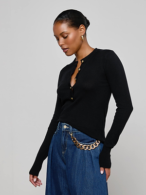A woman wears the L'AGENCE Ronda Knit Henley Top with lion head buttons and blue jeans with a gold chain belt, standing against a plain light background and looking down with her hand by her side.