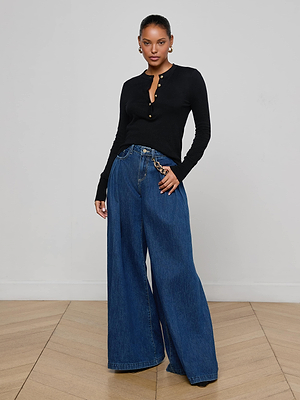 A woman poses indoors against a light background, wearing L'AGENCE's Ronda Knit Henley Top in black with lion head buttons, wide-leg blue jeans featuring a chain detail, hoop earrings, and her hair pulled back.