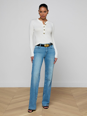 A woman stands confidently in a minimalist room wearing the Ronda Knit Henley Top in white, paired with light blue high-waisted jeans, a black belt with a gold buckle, and black heeled sandals.