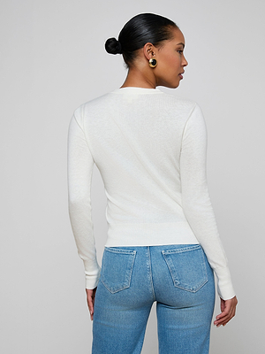 A woman with hair in a low bun, gold hoop earrings, and high-waisted blue jeans models the Ronda Knit Henley Top in white, featuring a fitted silhouette and button placket, standing against a plain background with her head turned sideways.