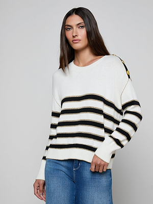 A woman with straight brown hair models the Bishop Striped Sweater, a relaxed fit knit with black horizontal stripes and gold buttons on one shoulder, paired with blue jeans against a plain light gray background.